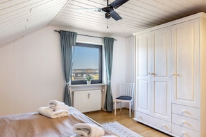 Une chambre à coucher comprenant un lit, une chaise, une armoire, une fenêtre avec des rideaux, un radiateur et un ventilateur de plafond.