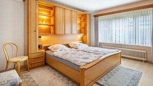 A bedroom with a wooden bed, built-in shelving, a chair, and a window with curtains. - Holiday Home 'Haus Lahrsen' with Private Terrace, Shared Garden and Wi-Fi (Wesselburen)