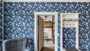 A room with a blue floral wallpaper, a grey armchair, a white bench with a hanging rack, and a door leading to another room with a painting. - Apartment 'Studio Zur »Alten Schmiede«' with Wi-Fi (Laage)