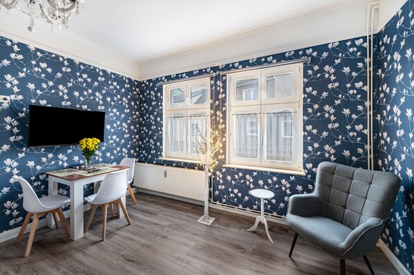 A room with a dining table, chairs, a TV, a window with white frames, and a patterned wall.