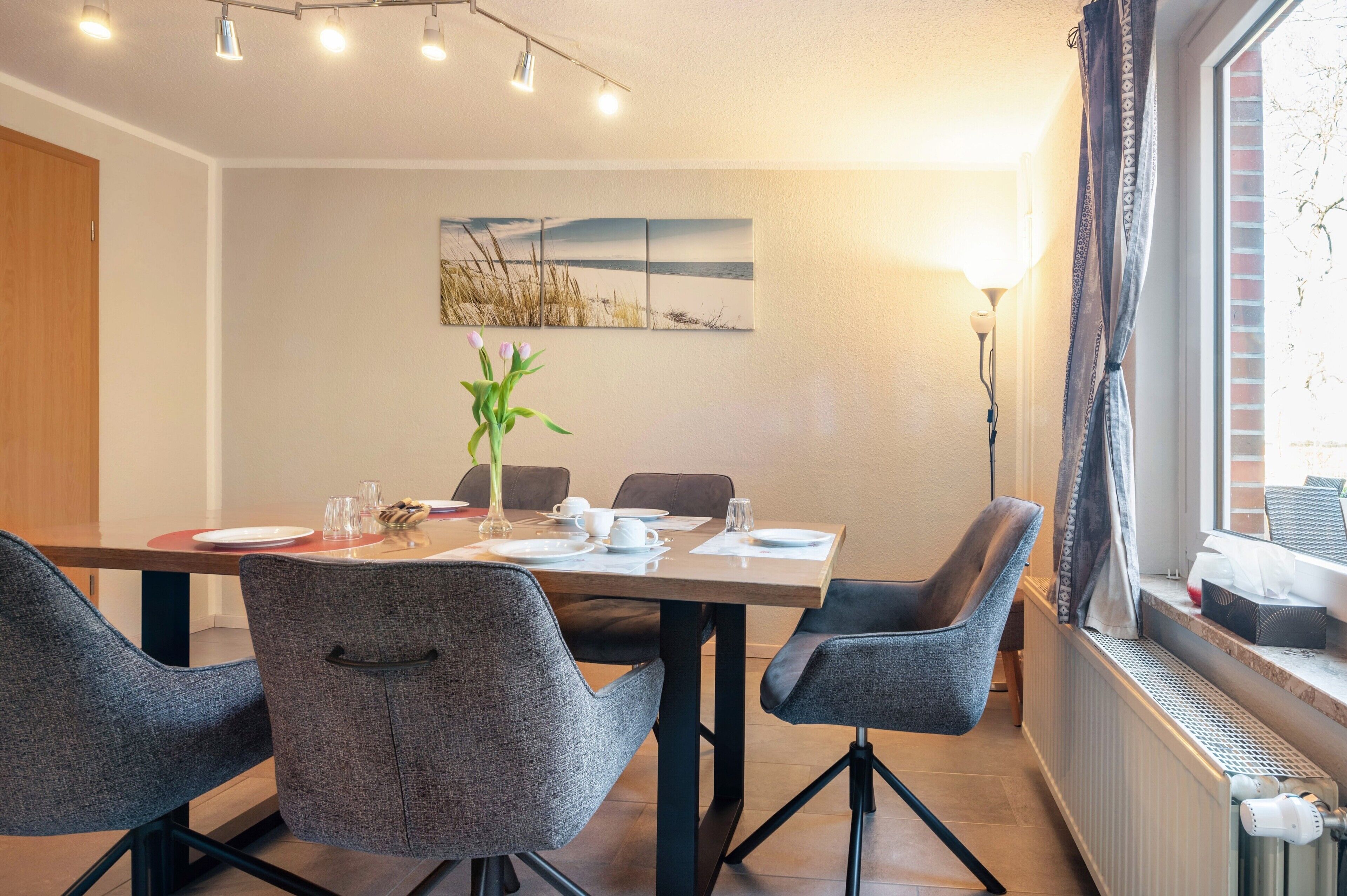 A dining area with a table set for four, chairs, a plant, and a painting on the wall.