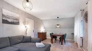 A modern living room with a grey sofa, a dining table with chairs, a wooden cabinet, and a wall-mounted clock. - Apartment 'Ferienwohnung Abendsonne' with Wi-Fi (Lehe)