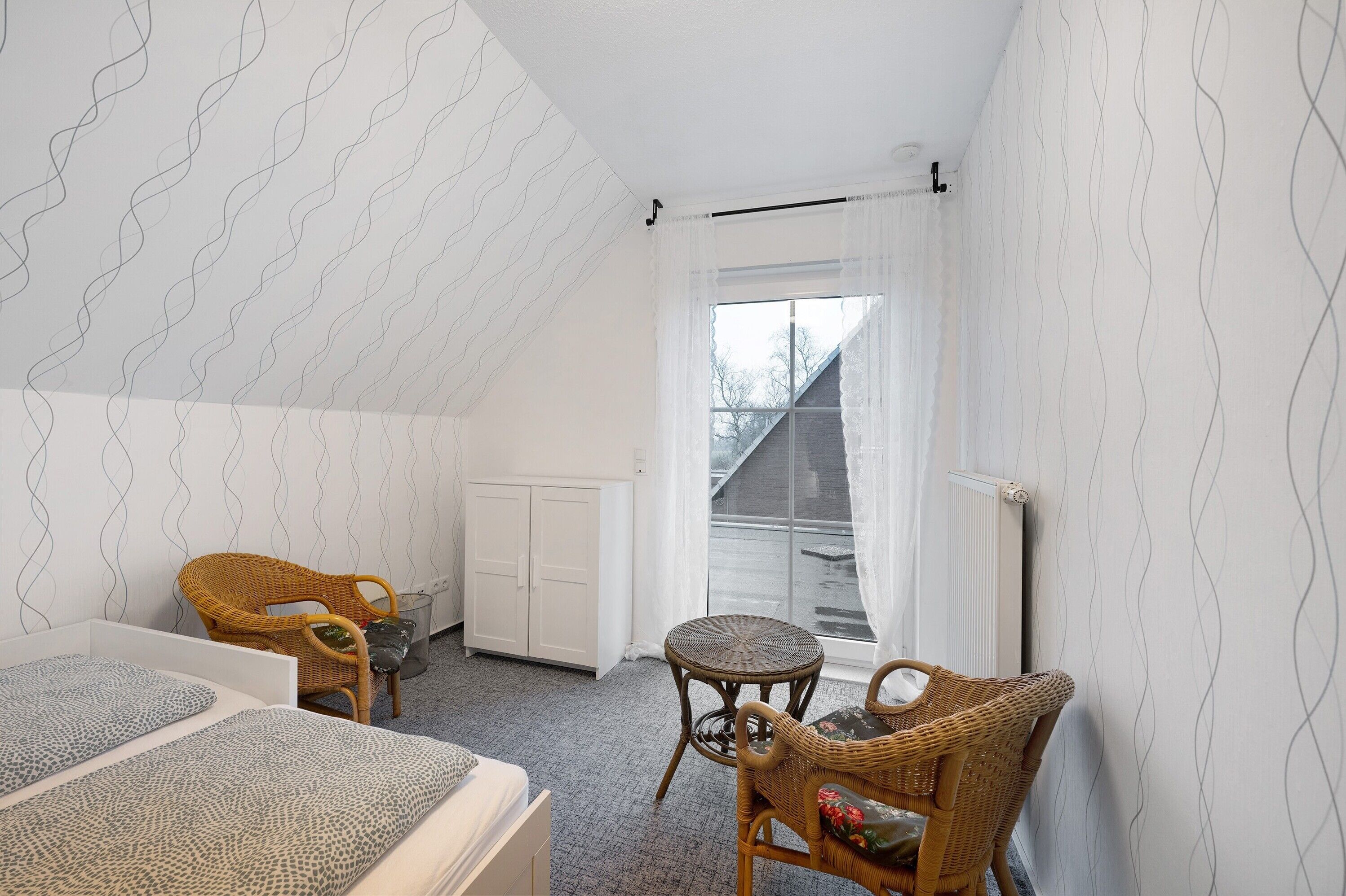 Un dormitorio con techo inclinado, una ventana con cortinas, una cama, dos sillas de mimbre y un radiador.