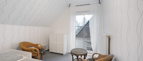 Un dormitorio con techo inclinado, una ventana con cortinas, una cama, dos sillas de mimbre y un radiador.