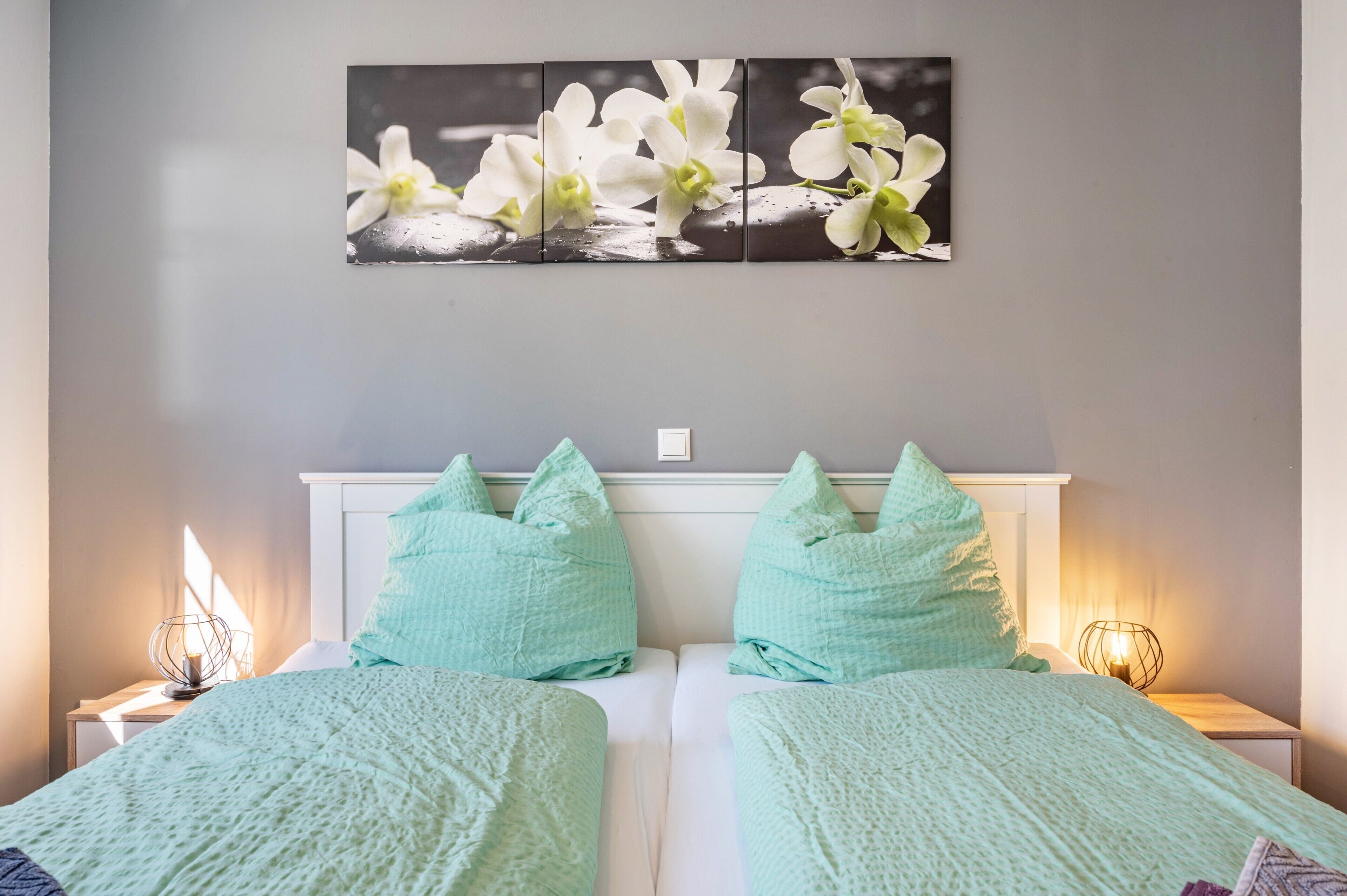 Une chambre avec deux lits, une literie de couleur turquoise, une œuvre d’art représentant une orchidée accrochée au mur et des lampes de chevet.