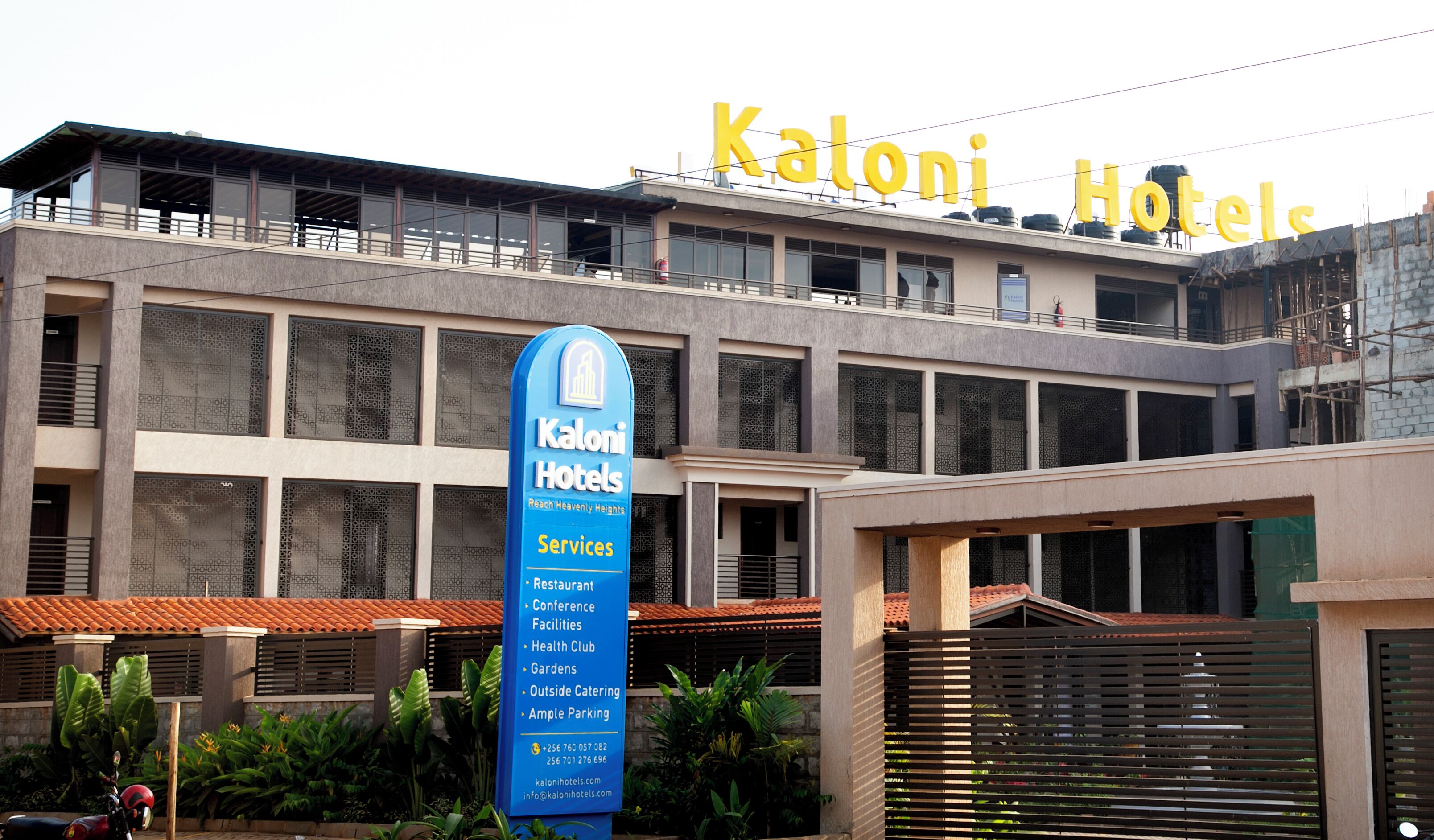 A hotel with a sign reading "Kaloni Hotels" and a blue sign listing services like restaurant, conference facilities, health club, and gardens.