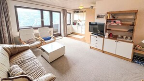 A living room with a sofa, armchair, coffee table, TV, and a kitchen in the background. - Daniele by Interhome (Mühlbach am Hochkönig)