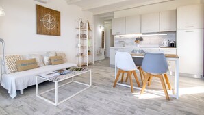 A modern living room with a sofa, coffee table, and a dining area with chairs. There is a kitchen in the background with white cabinets and a countertop. - Mare Azzurro by Interhome (Valledoria)
