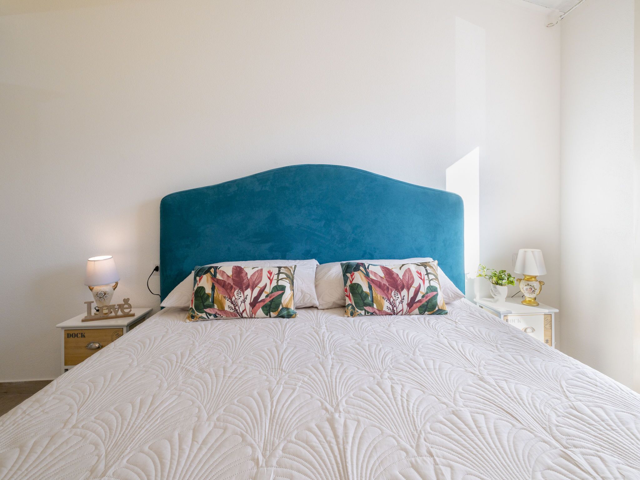 Een net opgemaakt bed met een blauw hoofdeinde, wit beddengoed en bloemenkussens. Er staat een nachtkastje met een lamp en een kleine plant.