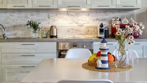 A modern kitchen with white cabinetry, a brick backsplash, and stainless steel appliances. There is a dining table with a decorative lighthouse centerpiece and a vase of flowers. - Onda perfetta by Interhome (Valledoria)