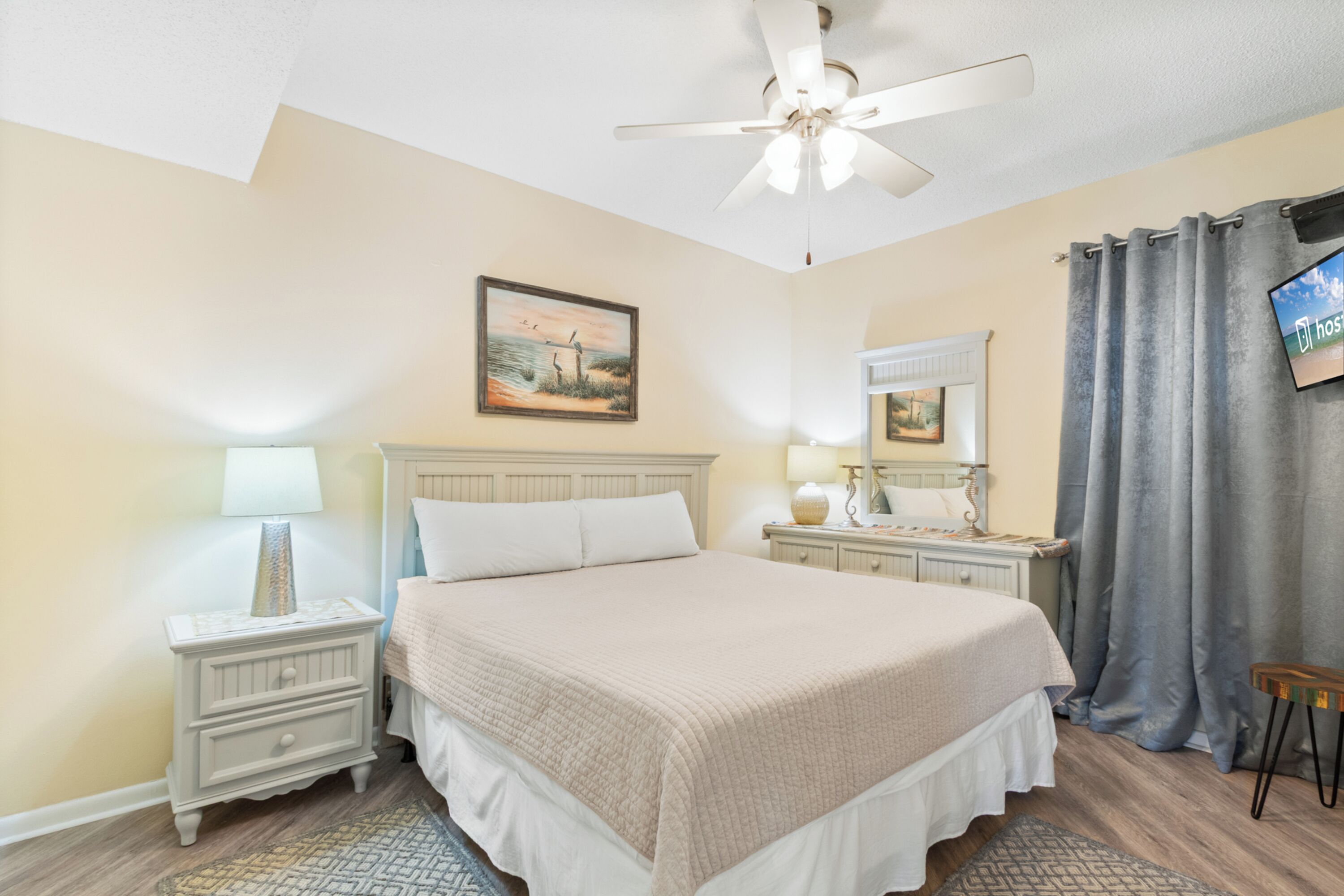 A bedroom with a bed, nightstands, a ceiling fan, a painting, a television, and a window with curtains.