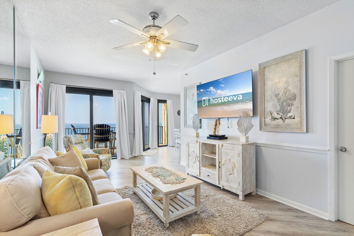 A modern living room with a sofa, coffee table, and artwork. A television is mounted on the wall, and there is a window with curtains.