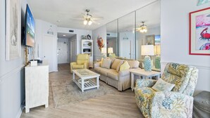 A spacious living room with a sofa, armchairs, and a coffee table. A ceiling fan is visible, and there is a large mirror on the wall. - Phoenix IV Condos (Orange Beach)
