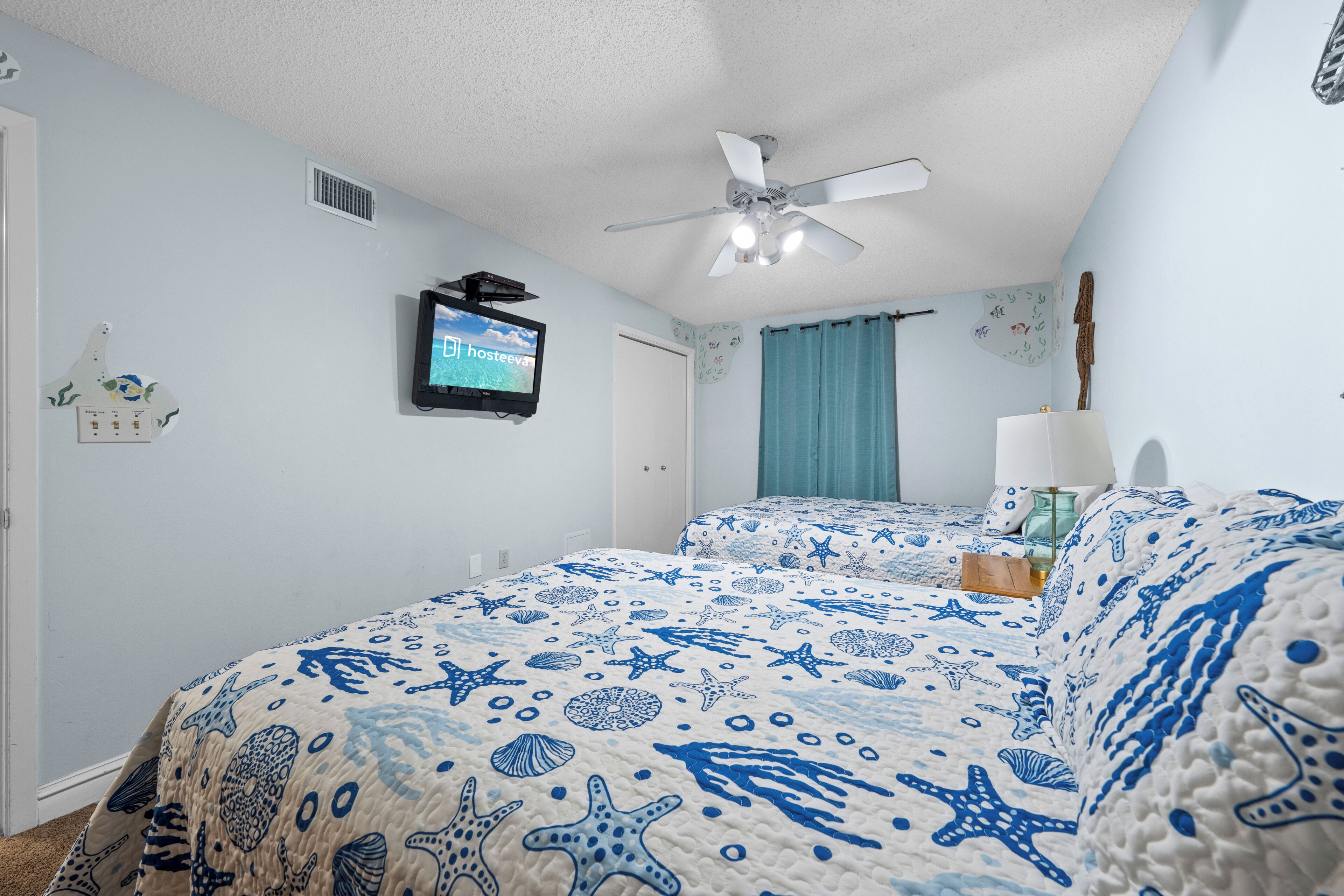 A bedroom with a bed featuring a blue and white nautical-themed bedspread, a ceiling fan, a mounted television, and a window with a blue curtain.