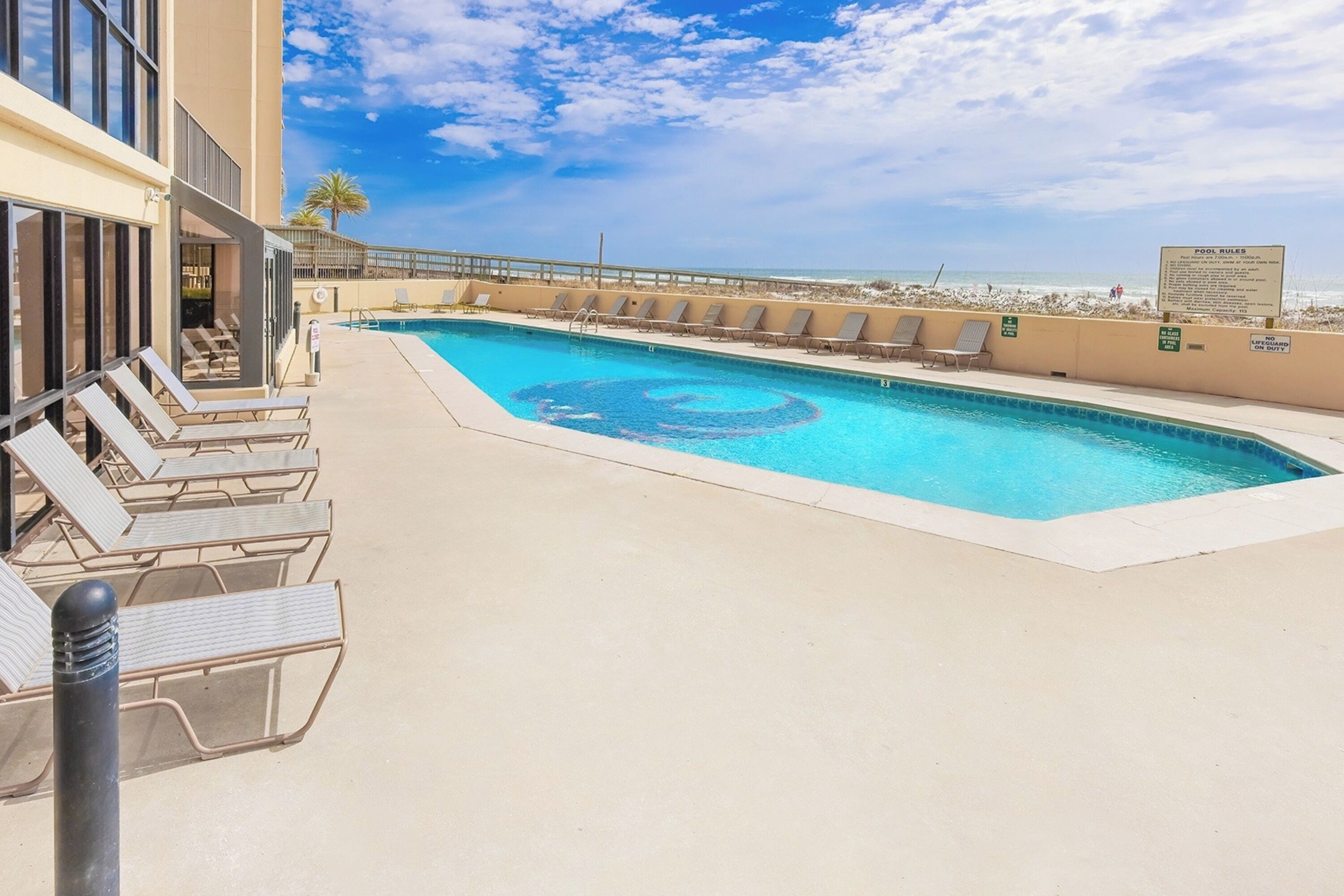 A swimming pool with lounge chairs and a lifeguard stand, surrounded by a building with a view of the beach.