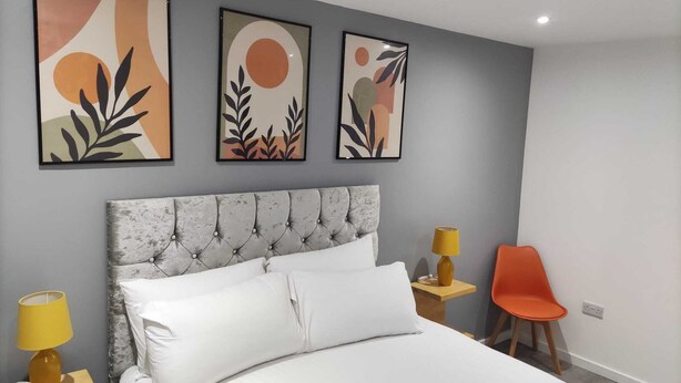 A bedroom with a tufted headboard, a bed with white linens, a nightstand with a lamp, and a chair. There are three abstract paintings on the wall.