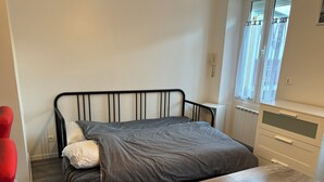 A bedroom with a bed, a wooden nightstand, a window with curtains, and a radiator. - Full center  (Le Malesherbois)