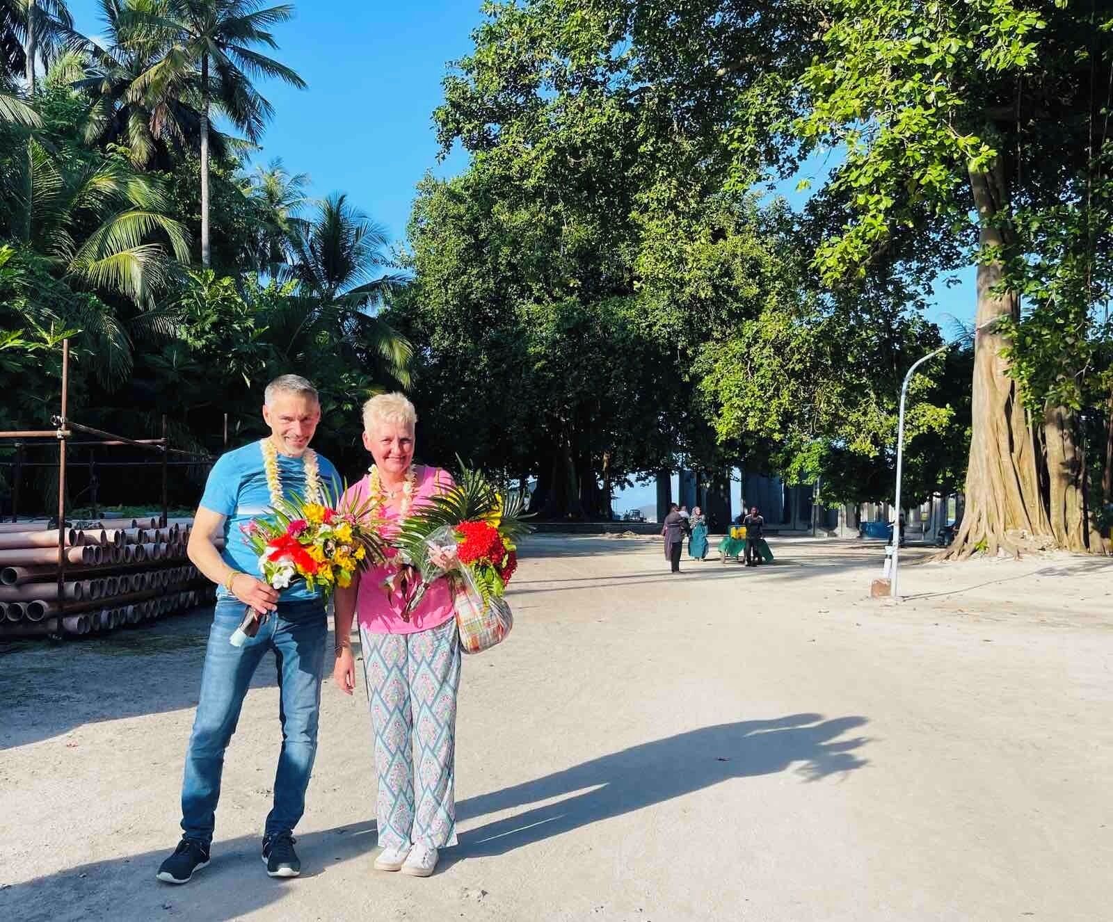 Duas pessoas segurando buquês de flores em um parque com árvores e um tronco de árvore grande ao fundo.