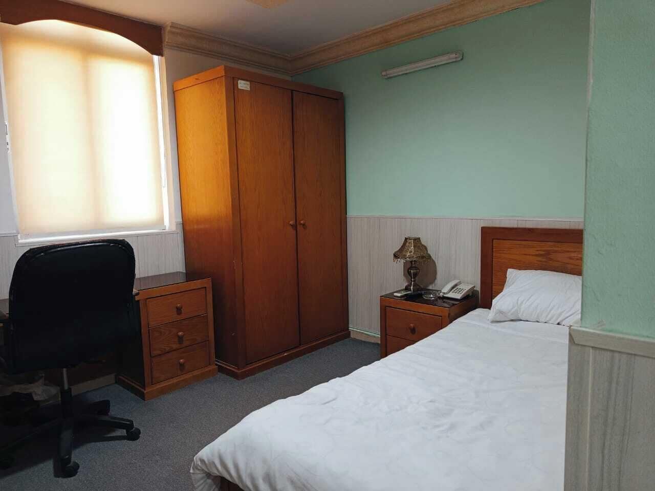A bedroom with a bed, a desk with a chair, a wardrobe, a bedside table with a lamp, and a telephone.