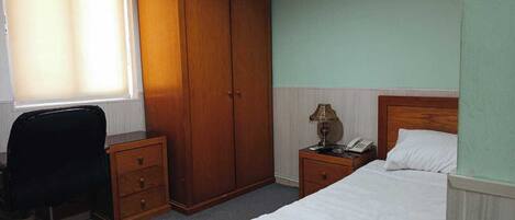 A bedroom with a bed, a desk with a chair, a wardrobe, a bedside table with a lamp, and a telephone.
