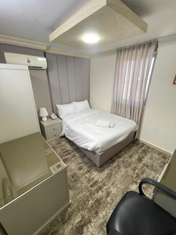 A hotel room with a bed, a desk, a chair, a nightstand, and an air conditioning unit.