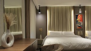 A modern hotel room with a large bed, a wooden desk, a chair, a vase with dried plants, and a painting on the wall. - Azmal Hotel & Suites (Irbid)