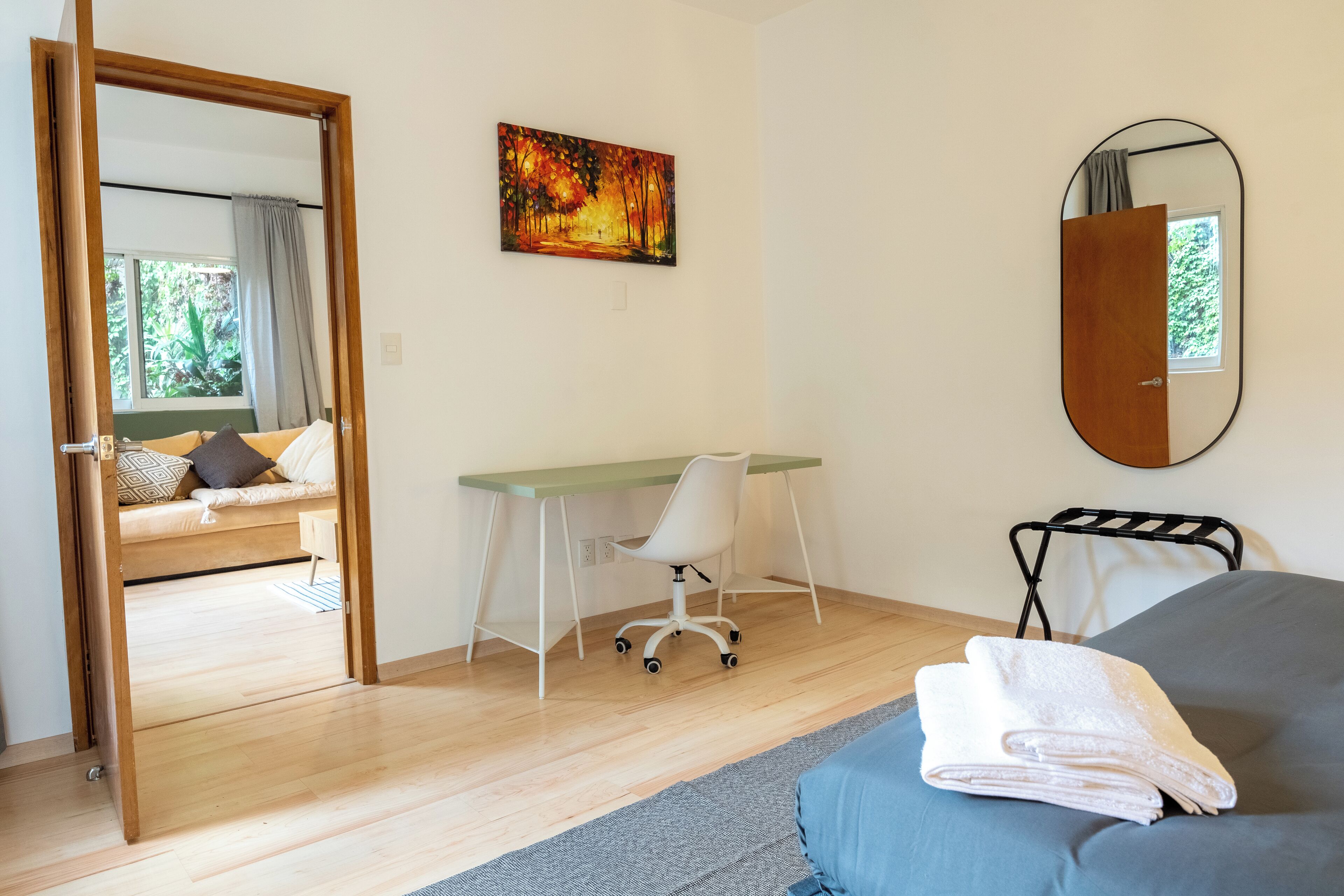 A bedroom with a bed, a desk, a chair, a mirror, and a painting on the wall.