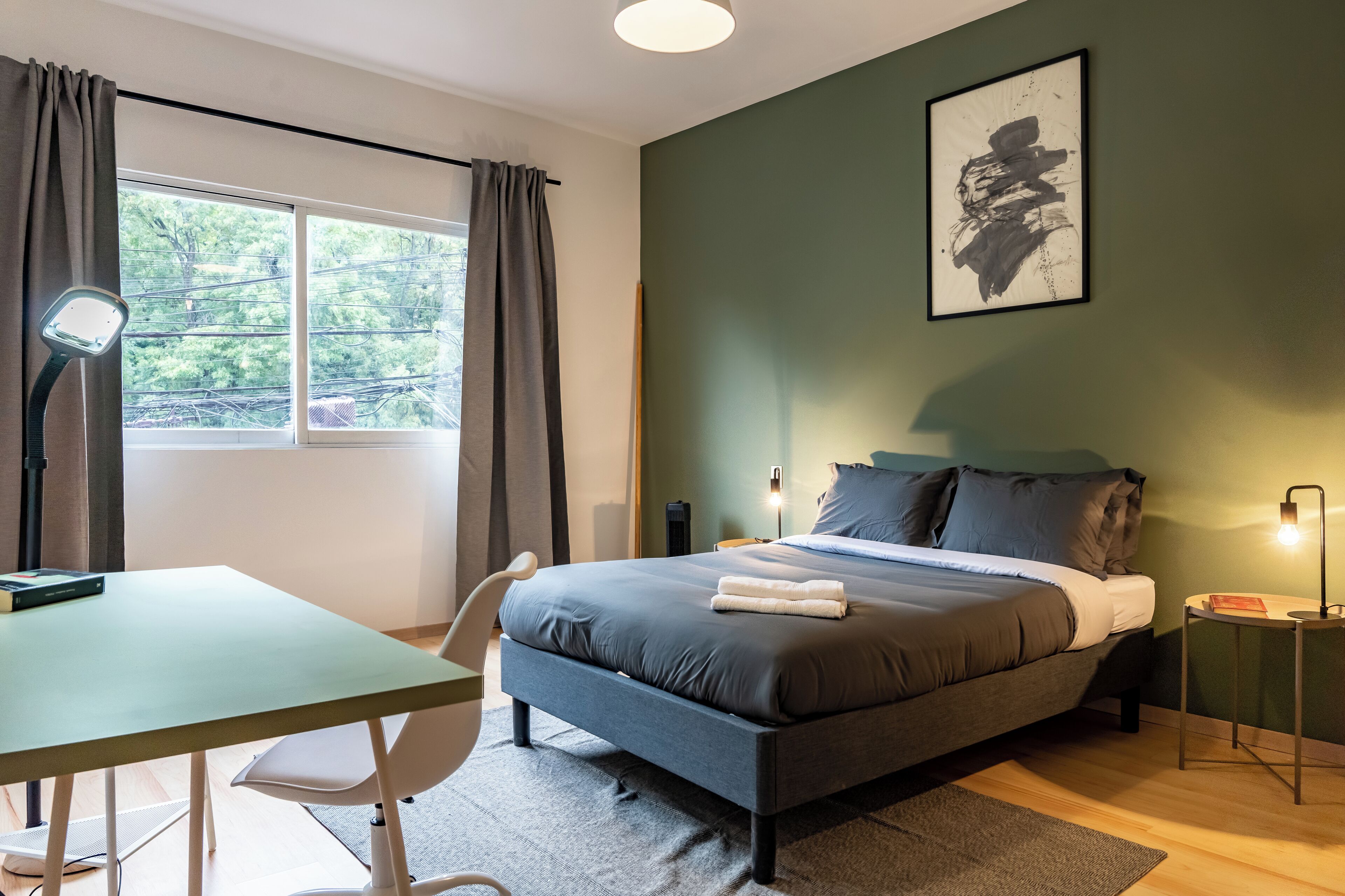 Ein Schlafzimmer mit Bett, Nachttisch, Lampe und Blick auf Bäume durch ein Fenster.