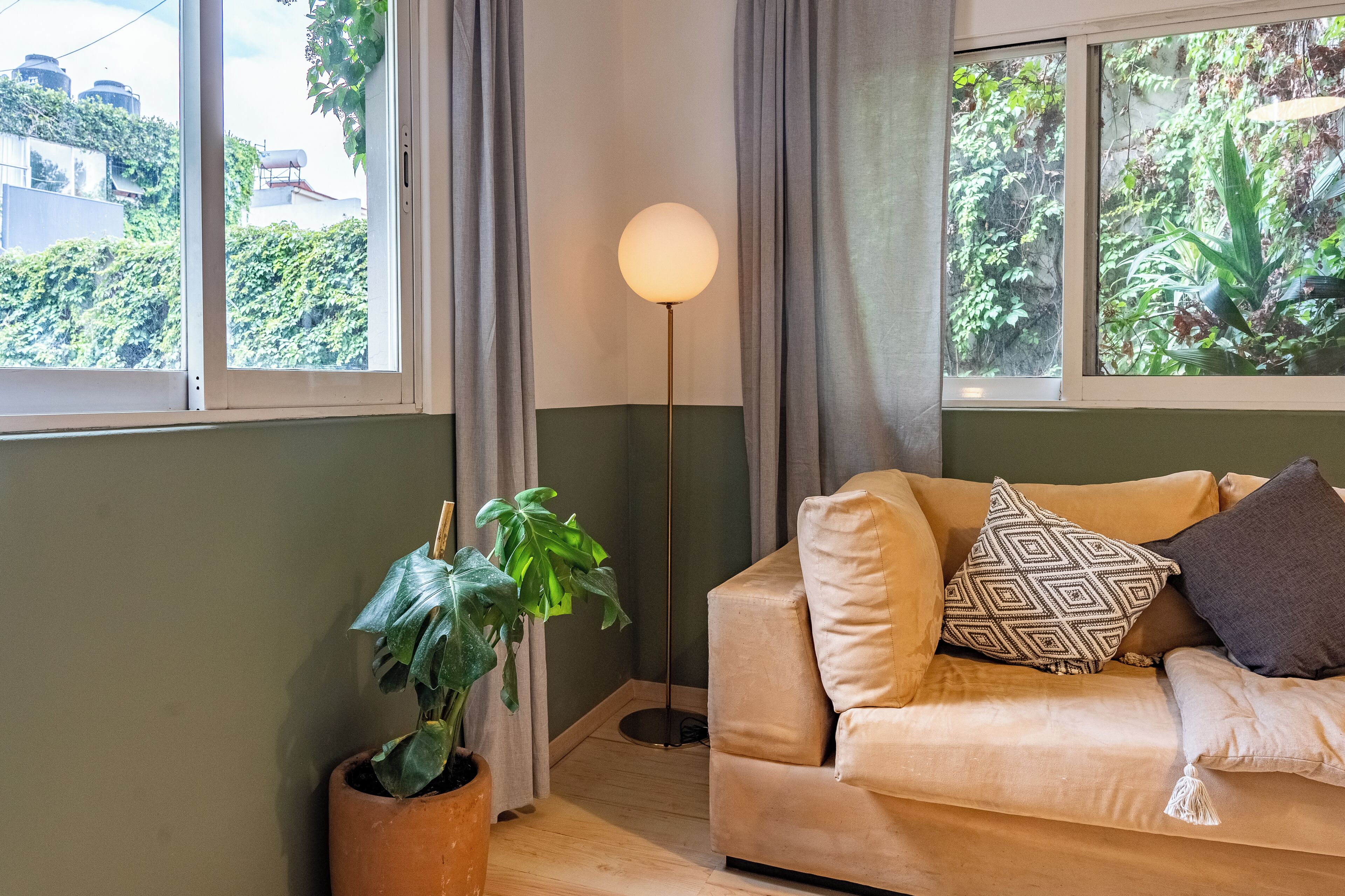 A room with a beige sofa, a floor lamp, a potted plant, and a view of greenery through the window.