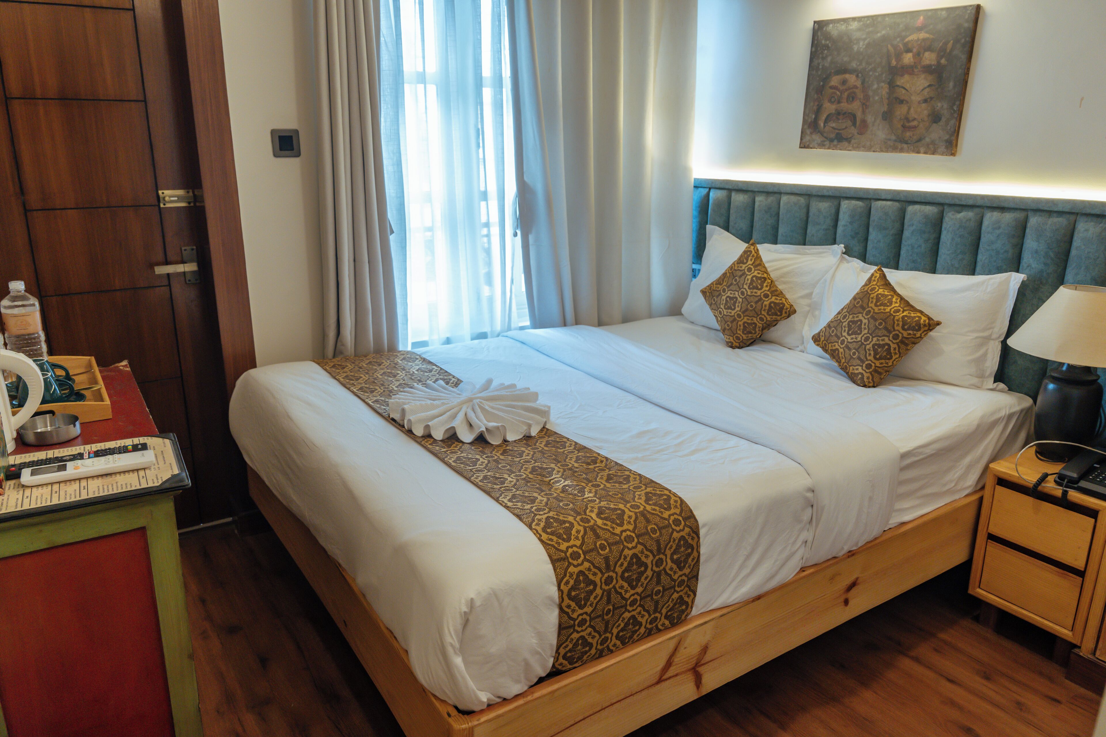 Een net opgemaakt bed met witte lakens en gestreepte kussens, een houten nachtkastje met een lamp, en een houten garderobe.