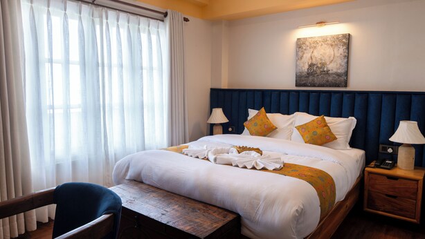 A hotel room with a large bed, bedside tables with lamps, a wooden bench, and a window with curtains. - Monkey Temple Residence (Nagarjun)