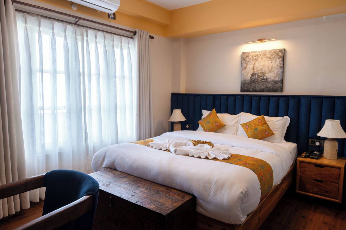 Habitación de hotel con una cama grande, mesitas de noche con lámparas, un banco de madera y una ventana con cortinas.