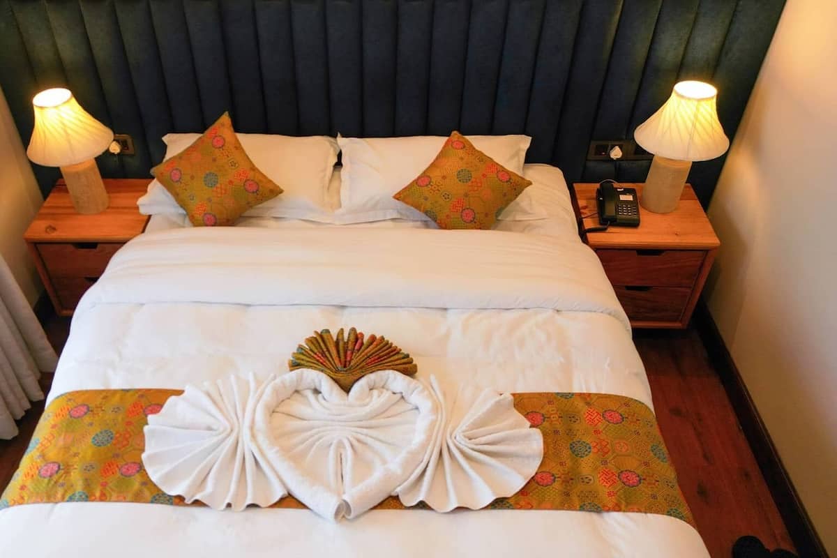 Una cama bien hecha con ropa blanca, dos almohadas decorativas y un arreglo de toallas con forma de corazón sobre la colcha.