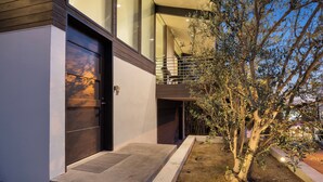 A modern two-story house with a large garage door, a balcony with a chair, and an olive tree in the yard.