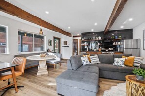 A modern living room with a grey sectional sofa, a round dining table, and a kitchen area with dark cabinetry and stainless steel appliances.