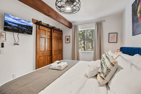 A bedroom with a bed, a large window, a wooden door, and a painting on the wall.