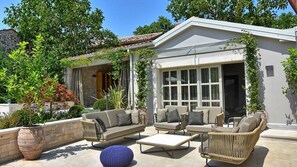 Uma área de estar externa com sofás e uma mesa de centro, cercada por plantas e um muro de pedra.