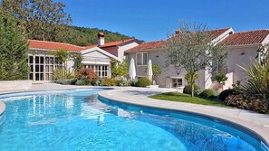 Uma piscina com borda curva, rodeada por um relvado bem cuidado e um edifício com telhado em telhas vermelhas.