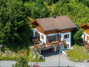 A two-story chalet with a balcony, surrounded by dense greenery.