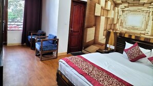 Quarto com cabeceira de madeira, cama com colcha vermelha e branca, mesa de cabeceira de madeira, uma cadeira e uma janela com cortinas.