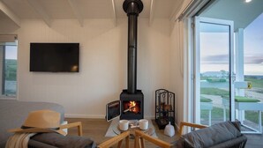 A cozy living room with a wood-burning stove, comfortable seating, a flat-screen TV, and a large window offering a scenic view.