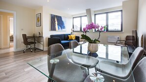 Un salon moderne avec une table à manger au plateau en verre, des chaises grises, un canapé bleu agrémenté de coussins jaunes et de grandes fenêtres laissant entrer la lumière naturelle.