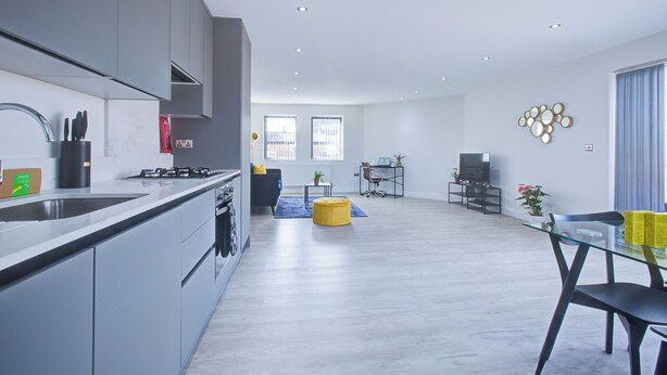 A modern kitchen with grey cabinets, a built-in stove, and a sink. There is a dining area with a glass table and chairs, a yellow ottoman, and a TV on a stand.