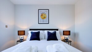 A neatly made double bed with white linens and two dark blue pillows, flanked by two bedside tables with lamps, in a room with a single framed artwork on the wall.