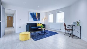 A modern living room with a blue sofa, a yellow ottoman, a coffee table, and a desk with a chair.