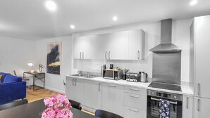 A modern kitchen with grey cabinets, a stainless steel range hood, and a dining area with a blue sofa and a table with pink flowers. - Hybrid Resi - Mitcham | 2 Bed Apartment (Mitcham)