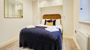Un dormitorio con un espejo grande, una cama con una colcha azul oscuro, almohadas blancas y un cabecero de madera.