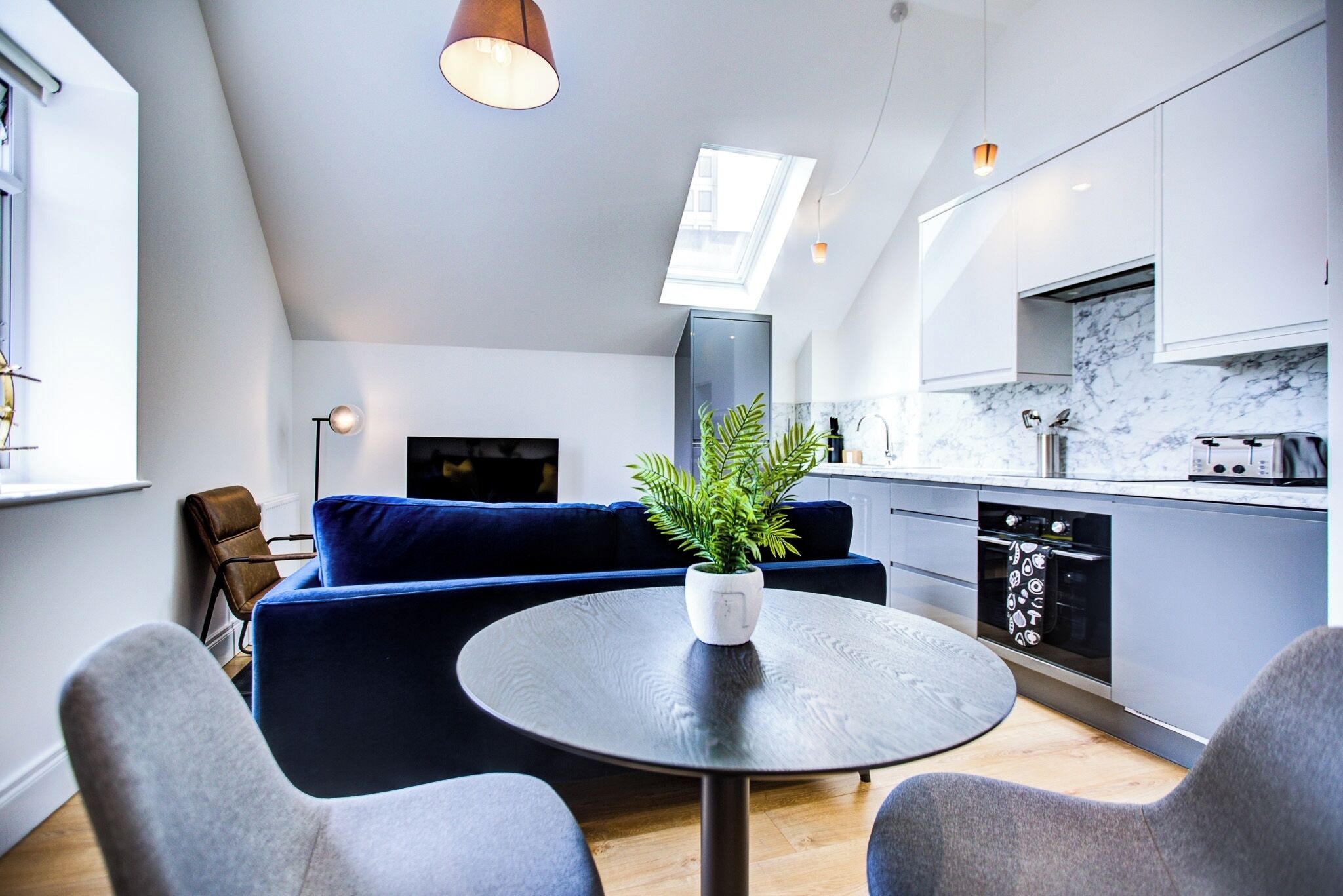 A modern kitchen with white cabinets, a marble backsplash, and a built-in oven. There is a blue sofa with a plant on it, and a round table with chairs in the foreground.