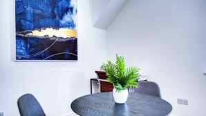 A modern dining area with a round table, two grey chairs, a potted plant, and a large abstract painting on the wall. - Hybrid Resi - New Malden | 1 Bed Apartment (New Malden)