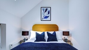 A neatly made bed with a dark blue blanket, two dark blue pillows, and a wooden headboard. There is a framed artwork above the bed, and two bedside lamps on either side. - Hybrid Resi - New Malden | 1 Bed Apartment (New Malden)